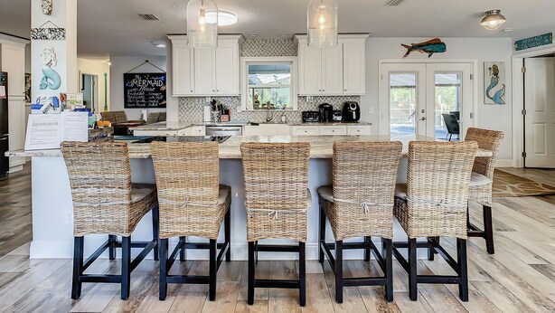 A modern kitchen with white cabinets, a central island with wicker chairs, pendant lighting, and decorative wall art.