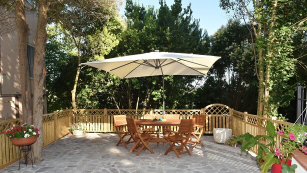 An outdoor patio with a stone floor, wooden table and chairs, a large umbrella, and potted plants. - Appartamento in zona Panoramica a due passi dal Mare ed immerso nel verde (Gabicce Monte)