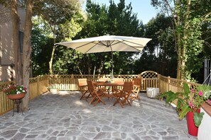 Une terrasse extérieure avec un sol en pierre, une table et des chaises en bois, un grand parasol et des plantes en pot.