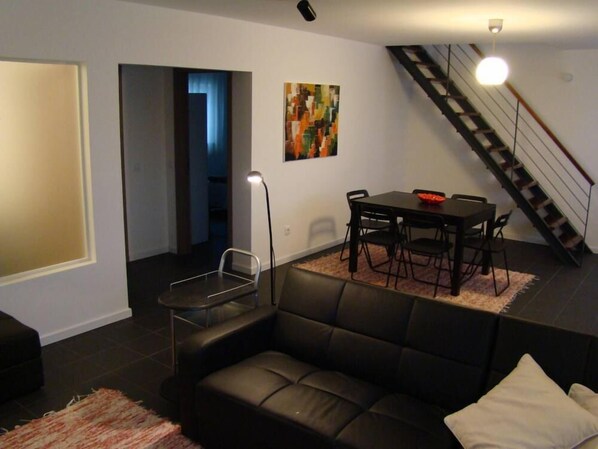 A modern living room with a black leather sofa, a dining table with chairs, a staircase, and a painting on the wall.