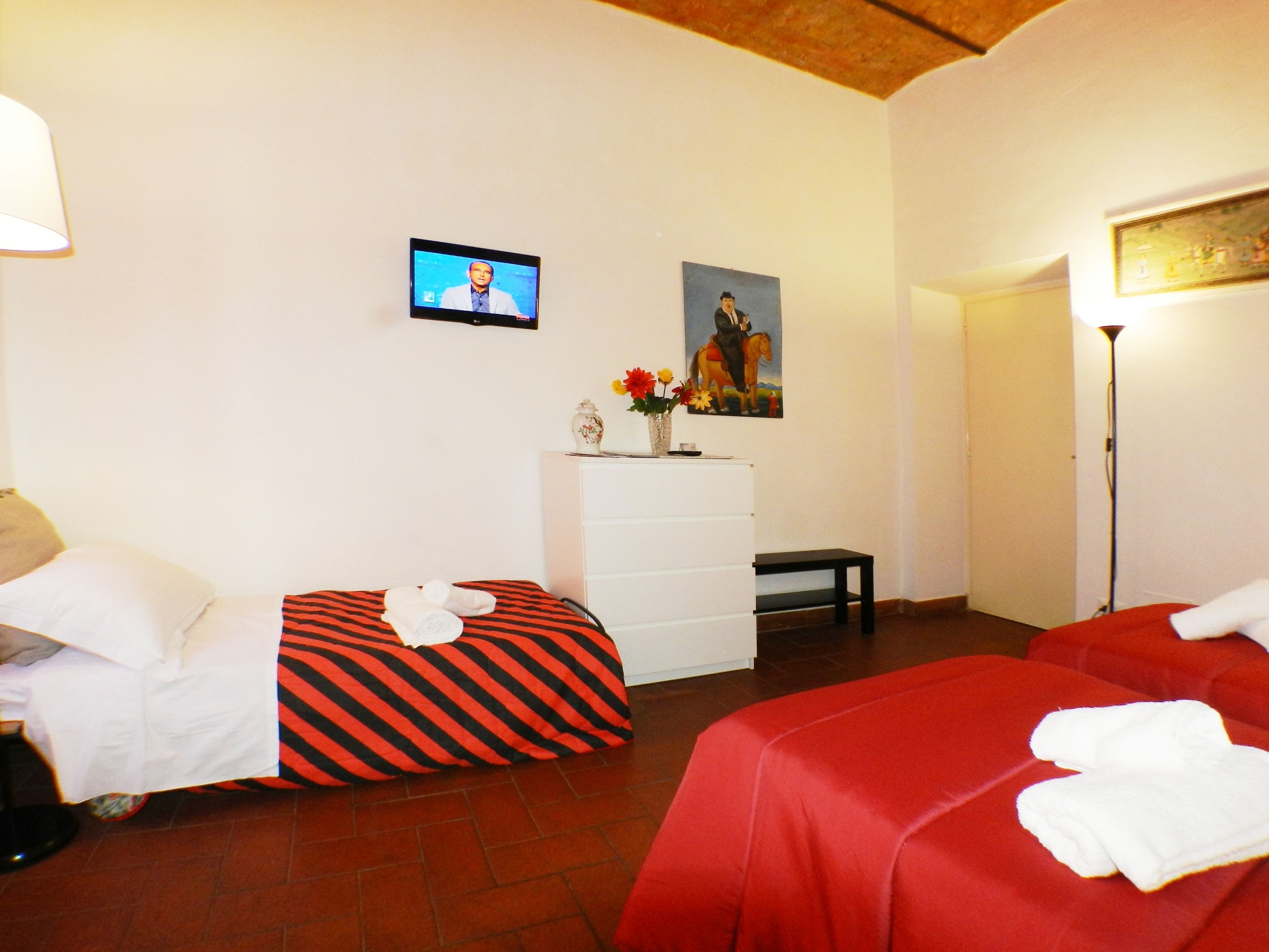 Habitación de hotel con dos camas, un televisor en la pared, un aparador, un cuadro y un florero con flores.