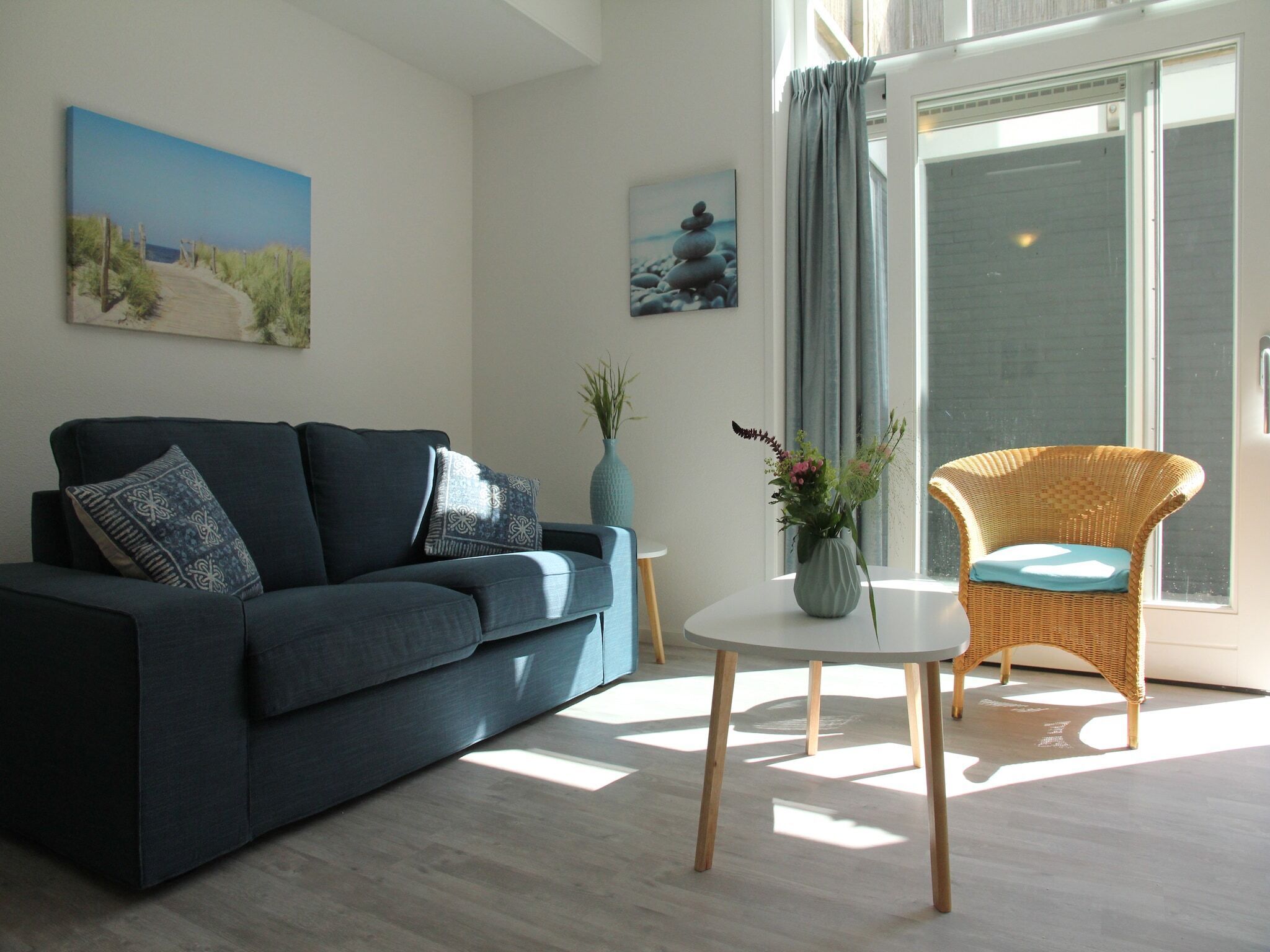 A modern living room with a dark blue sofa, a wicker chair, a small round table, and two paintings on the walls.