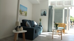 Un salon moderne avec un canapé bleu foncé, une petite table ronde surmontée d’une lampe et un fauteuil en osier. Deux tableaux ornent le mur, une fenêtre est agrémentée de rideaux et offre une vue sur l’extérieur.