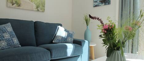 A living room with a blue sofa, a white round table, and a vase with flowers. There are two paintings on the wall.