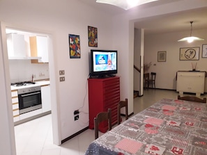 Ein Wohnzimmer mit Fernseher, einem Esstisch mit gemustertem Tischläufer und einer Küche im Hintergrund.