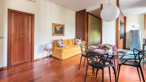 A modern living room with a wooden floor, a yellow sofa, a dining table with chairs, and a hanging light fixture.