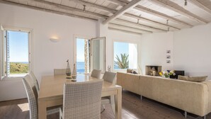 Habitación amplia con una mesa de comedor de madera, sillas de mimbre, un sofá y un televisor. Grandes ventanas ofrecen vistas al exterior.