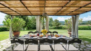 Un espacio de comedor al aire libre con mesa y sillas cubiertas, y vistas a un jardín exuberante y colinas al fondo.