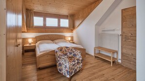A bedroom with a wooden ceiling, a bed with white linens, a floral-patterned chair, and a wooden bench. - Sunny apartment in an ideal location for active people, families, Genießer (Kaltern)