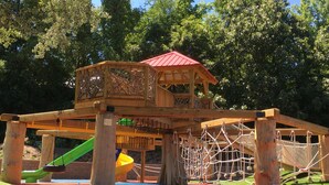 A wooden play structure with a red roof, a slide, and a rope net. - Downtown Main Street "STONES" FENCED YARD (Blowing Rock)
