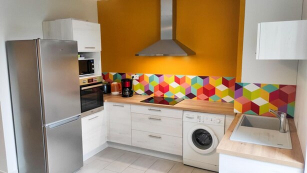 Una cucina compatta con frigorifero in acciaio inossidabile, forno a microonde, macchina da caffè, lavatrice e un vivace rivestimento geometrico.