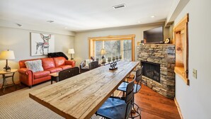 A spacious living room with a long wooden table, leather sofa, and a stone fireplace.
