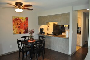 A dining area with a table and chairs, a ceiling fan, a kitchen with a refrigerator, and a painting on the wall.