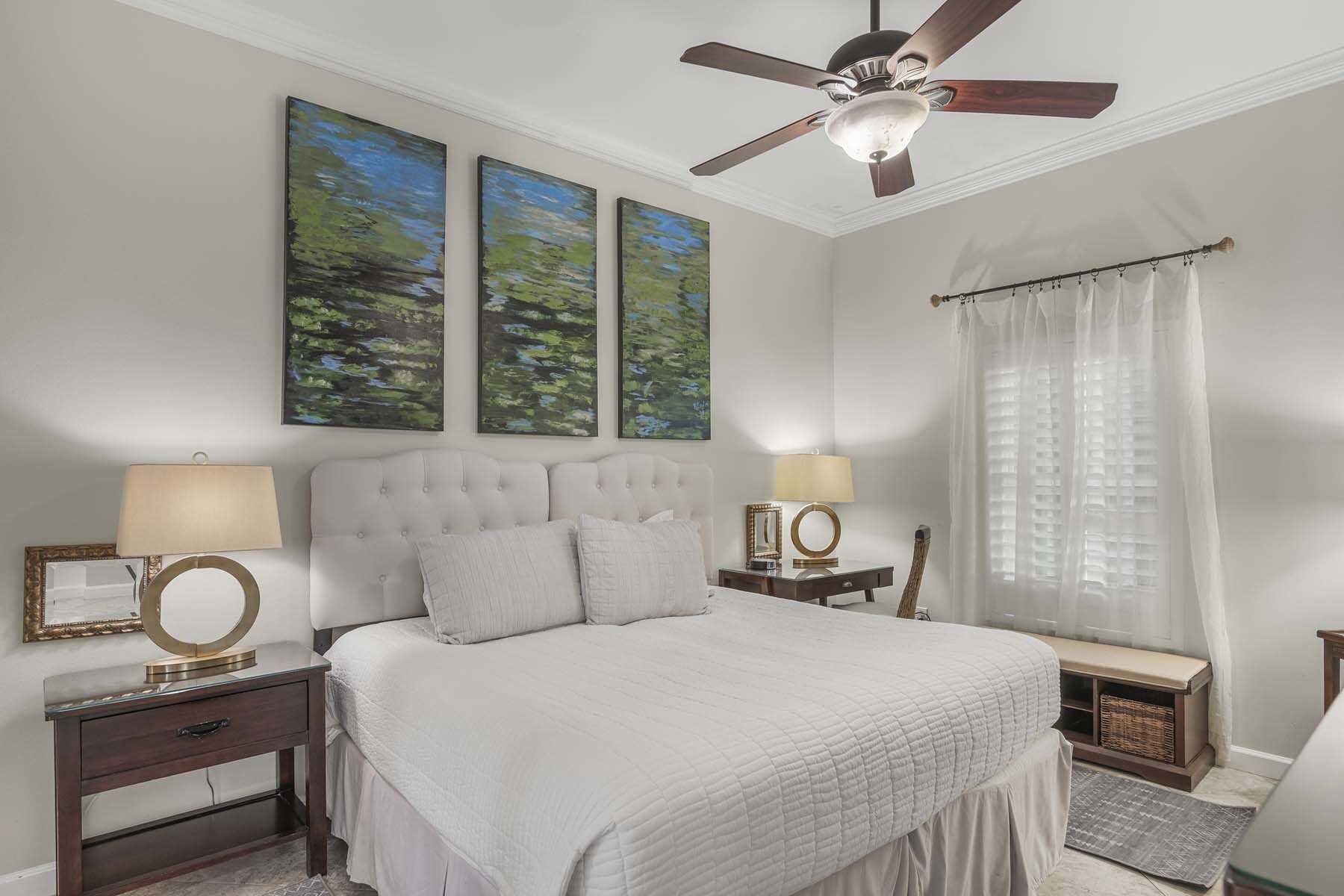 A bedroom with a large bed, two bedside tables with lamps, a ceiling fan, and two abstract paintings on the wall.