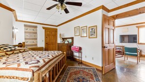 A bedroom with a large bed, wooden headboard, a ceiling fan, a wooden door, a rocking chair, and a television in the adjacent room. - Comfy Country Condo (West) on 10 Acres between Bozeman & Big Sky (Gallatin Gateway)
