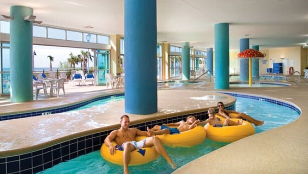 Ein Hallenbad mit Personen auf Luftmatratzen, großen blauen Säulen und Blick auf den Ozean durch große Fenster.