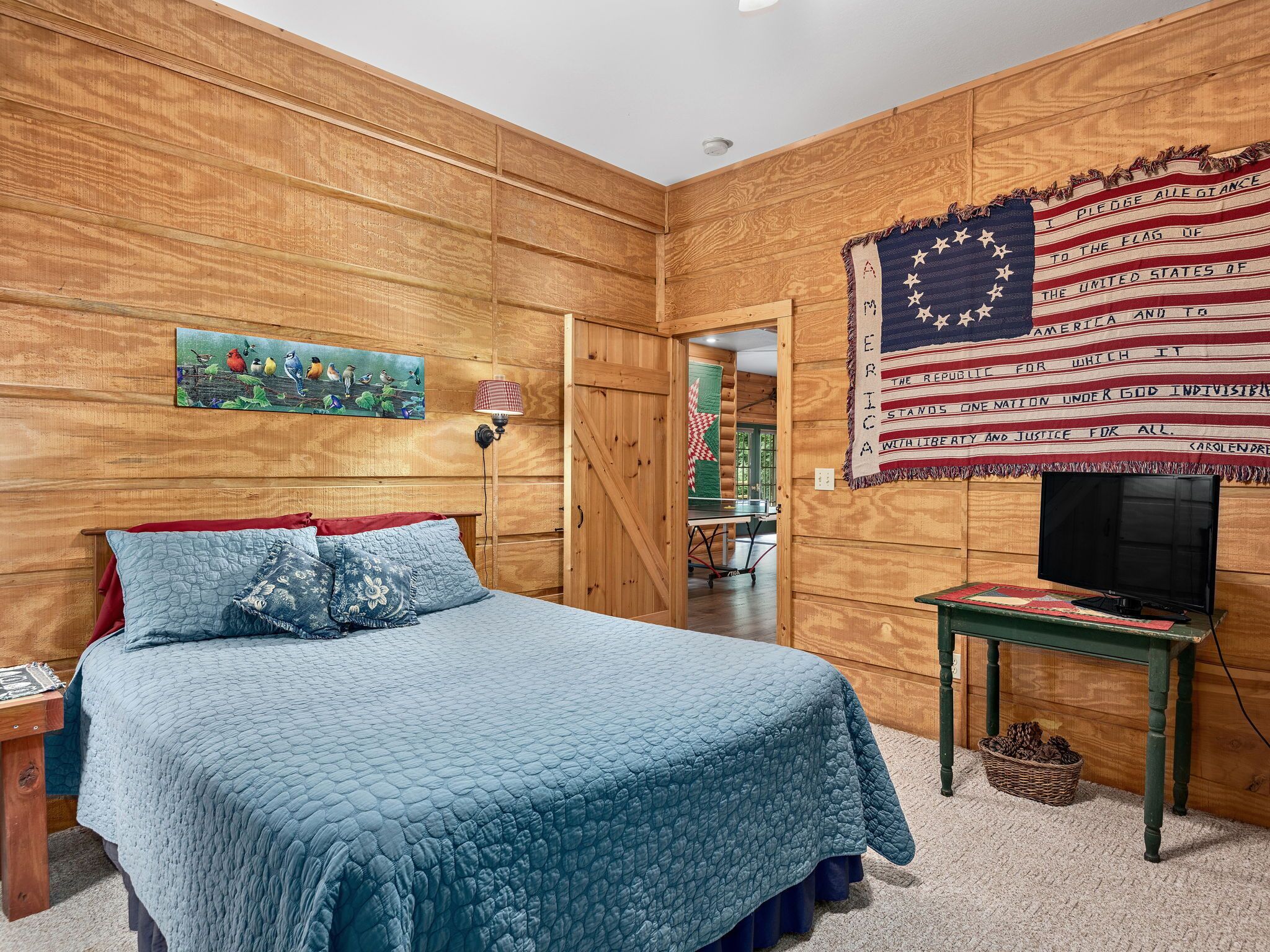 Une chambre avec un mur recouvert de panneaux de bois, un lit avec une literie bleue, un téléviseur posé sur un meuble en bois et un drapeau américain.