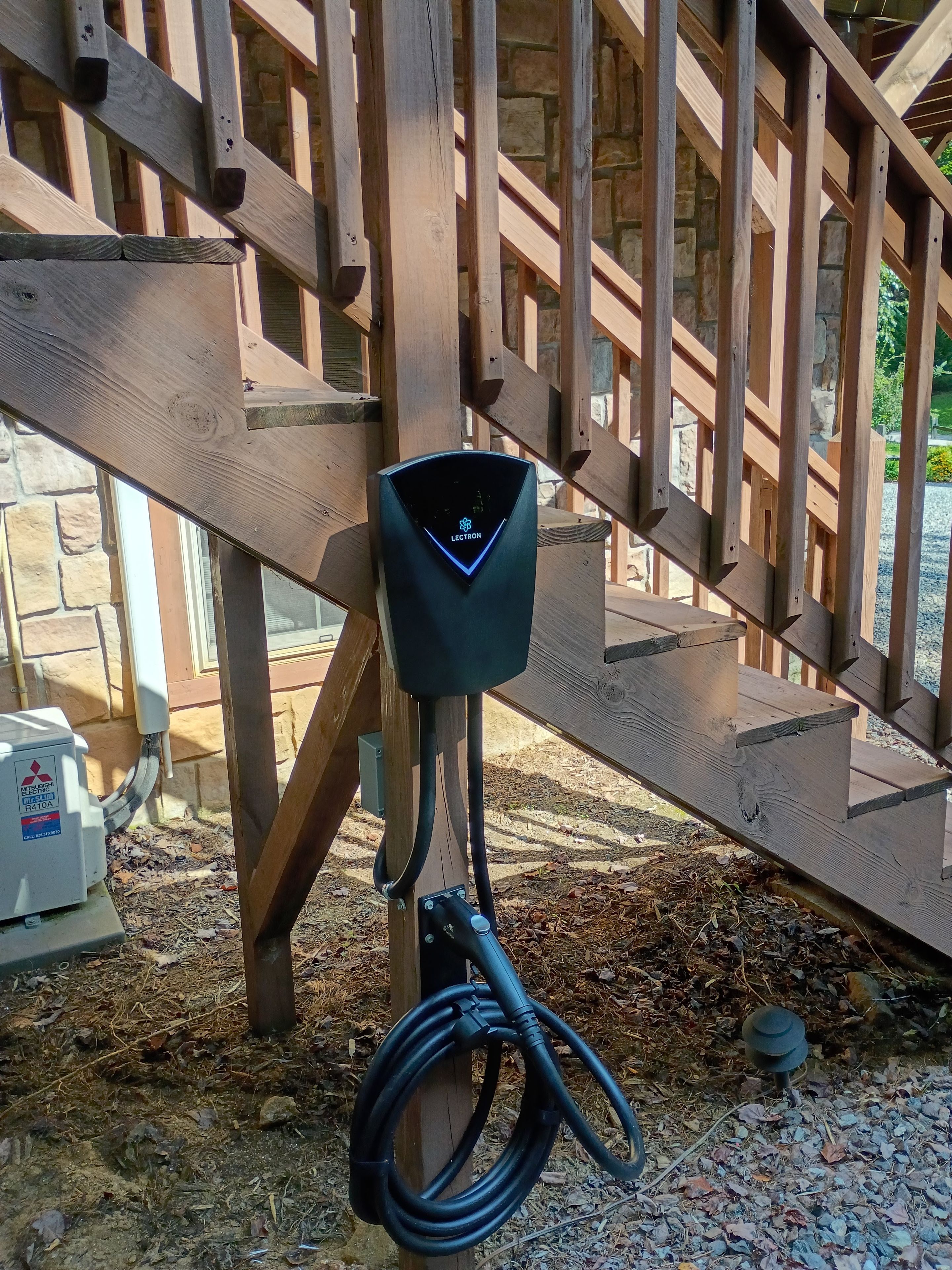 A black electric vehicle charging station attached to a wooden structure with a stone wall in the background.