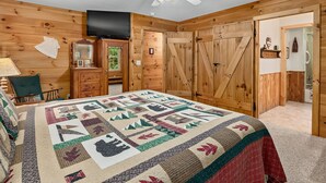 Une chambre confortable avec un grand lit, des murs en bois, un ventilateur de plafond, une télévision et une porte donnant sur une salle de bain.