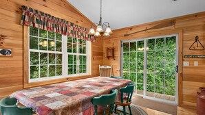 L’intérieur d’un chalet en bois comprend une table à manger, des chaises, un lustre et de grandes fenêtres donnant sur un paysage verdoyant.