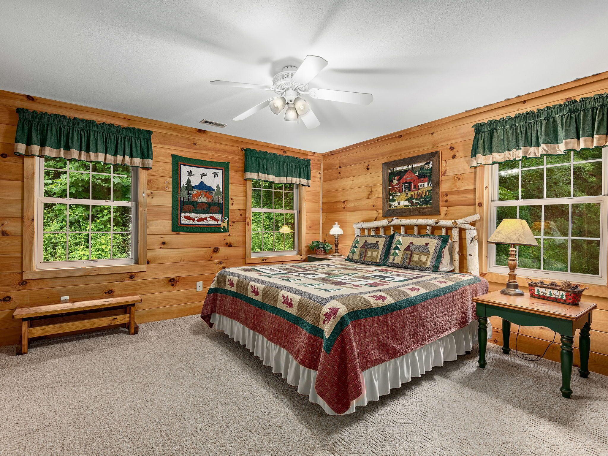 A cozy log cabin bedroom with a large bed, a ceiling fan, and two windows with curtains.