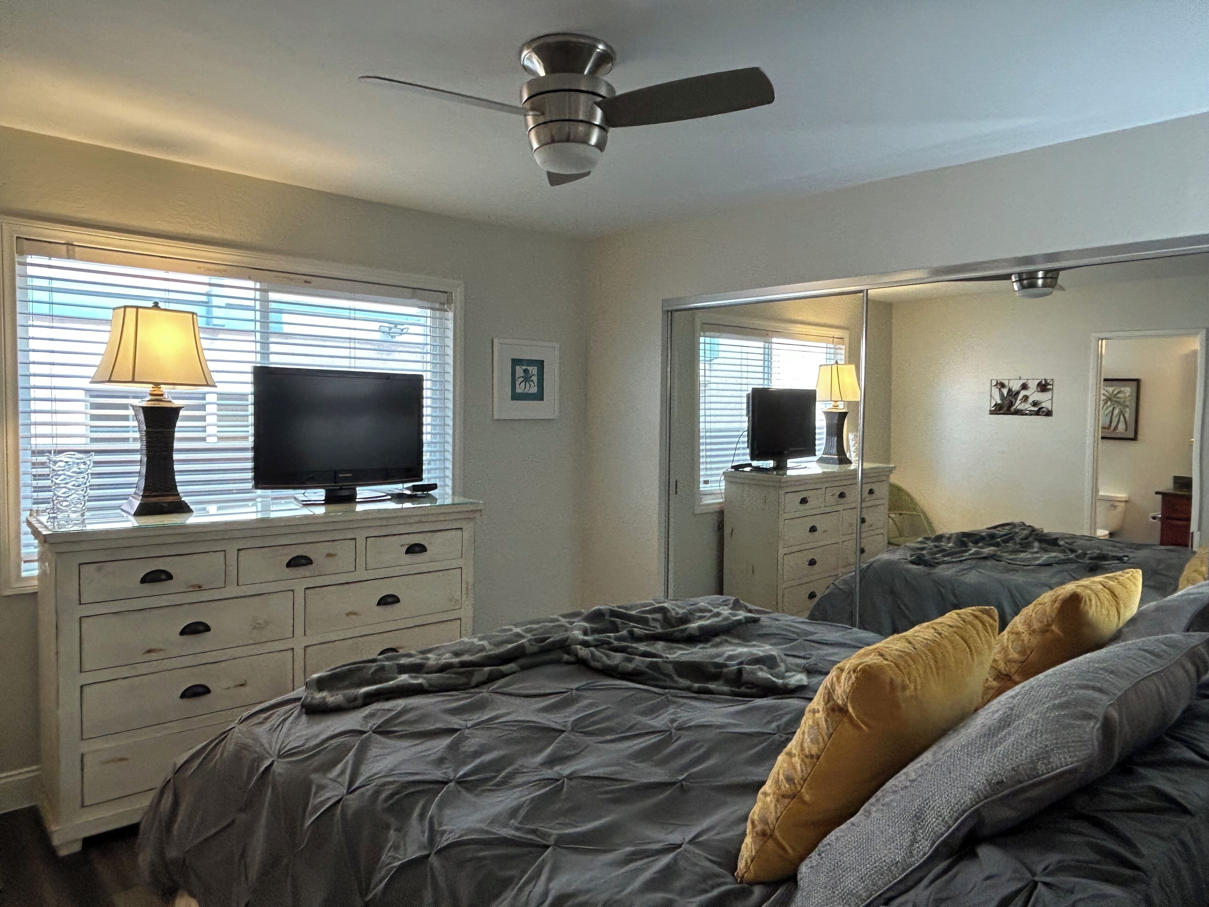 A bedroom with a bed, a TV on a dresser, a ceiling fan, and a window with blinds.