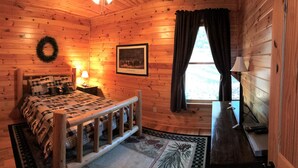 A cozy log cabin bedroom with a bed, a nightstand, a lamp, a framed picture, and a window with curtains. - Near Wine Country & Parks. Great Hiking & Amazing Views! Reduced June rates! (Hays)