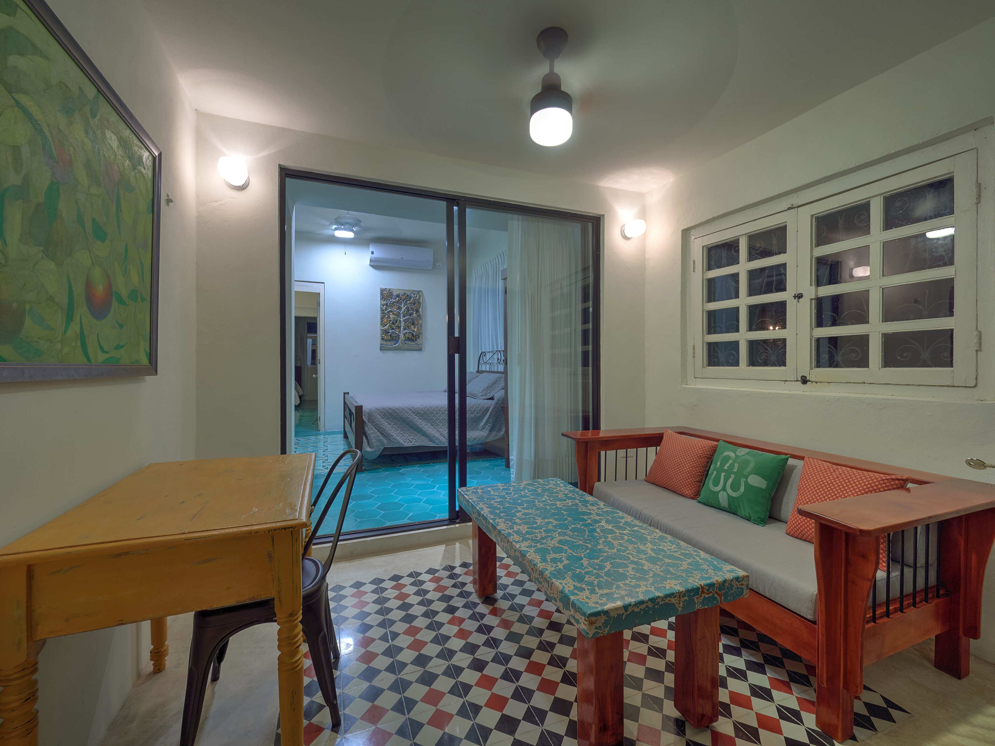 A small, well-lit room with a bed, a table, and a bench. There is a window with multiple panes and a door leading to another room.