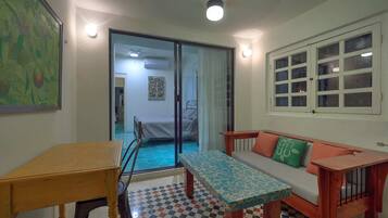 A small, well-lit room with a bed, a table, and a bench. There is a window with multiple panes and a door leading to another room.