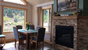 Un comedor con mesa y sillas, una chimenea y ventanas amplias con vistas a un paisaje boscoso.