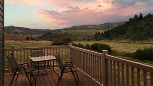 Una terraza de madera con una mesa y sillas, con vistas a un paisaje montañoso bajo un cielo al atardecer.