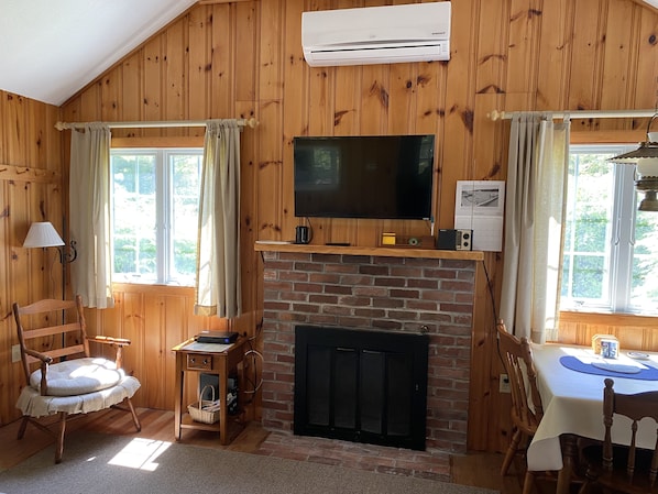 A cozy room with wooden walls, a fireplace, a television, and a dining table. - Brewster, Beach Cottage with 1/2 Mile of Private Asso. Beach for your Enjoyment. (Brewster)