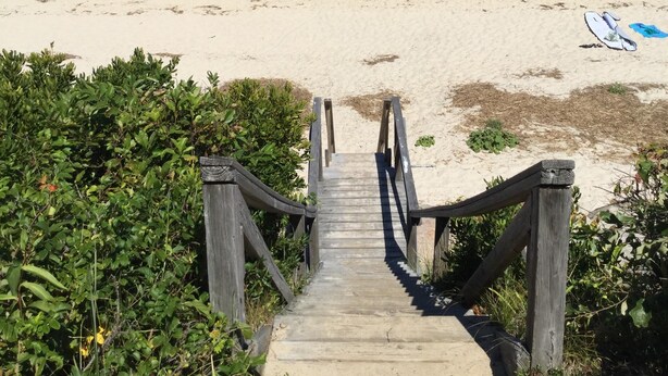 Un sendero de madera que lleva a una playa con barcos en el agua y un cielo despejado.