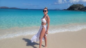 Una mujer de pie en una playa arenosa con agua turquesa cristalina y una pequeña isla al fondo.