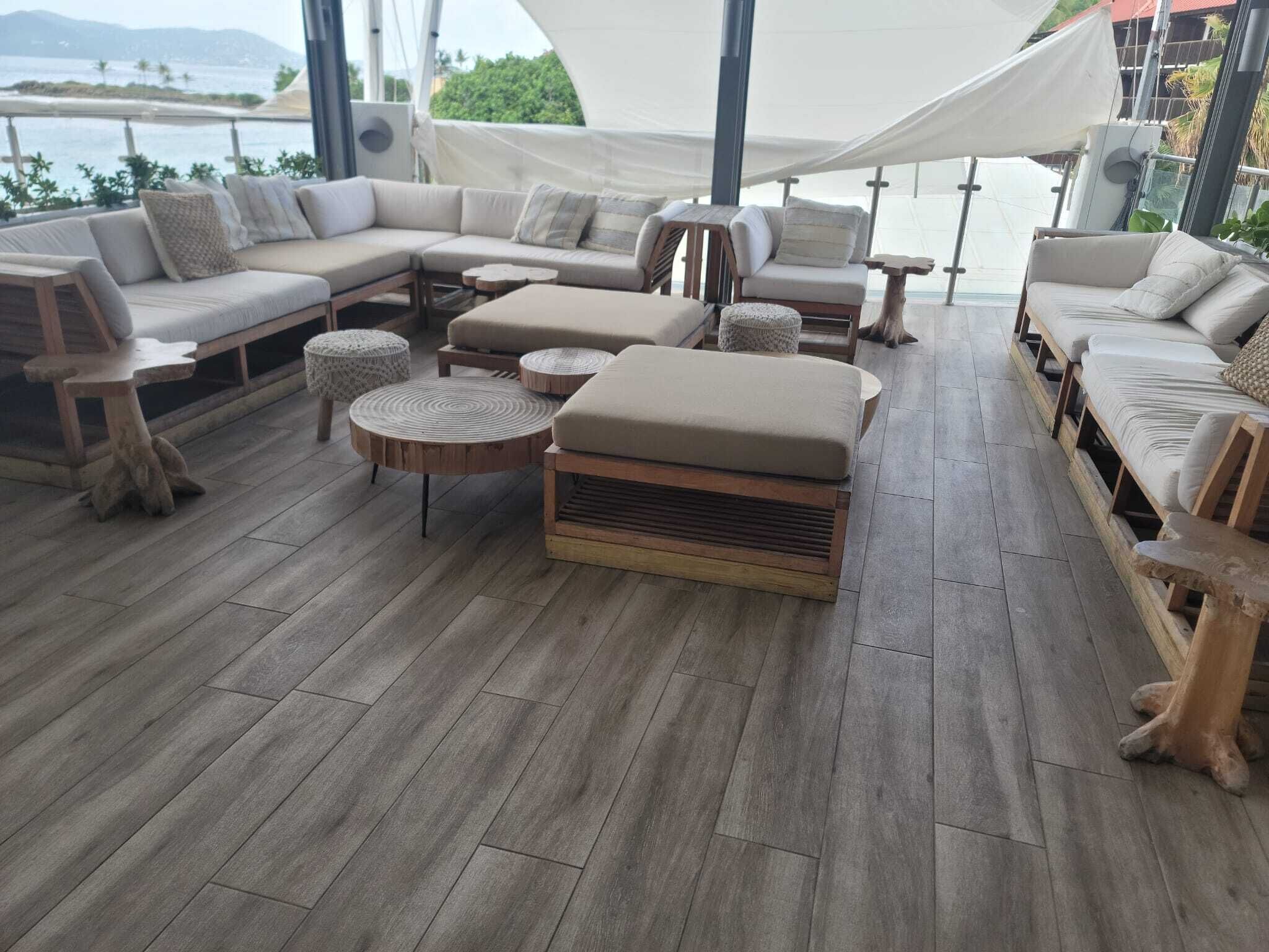 A spacious outdoor seating area with a large sofa, ottoman, and round tables, overlooking a body of water.
