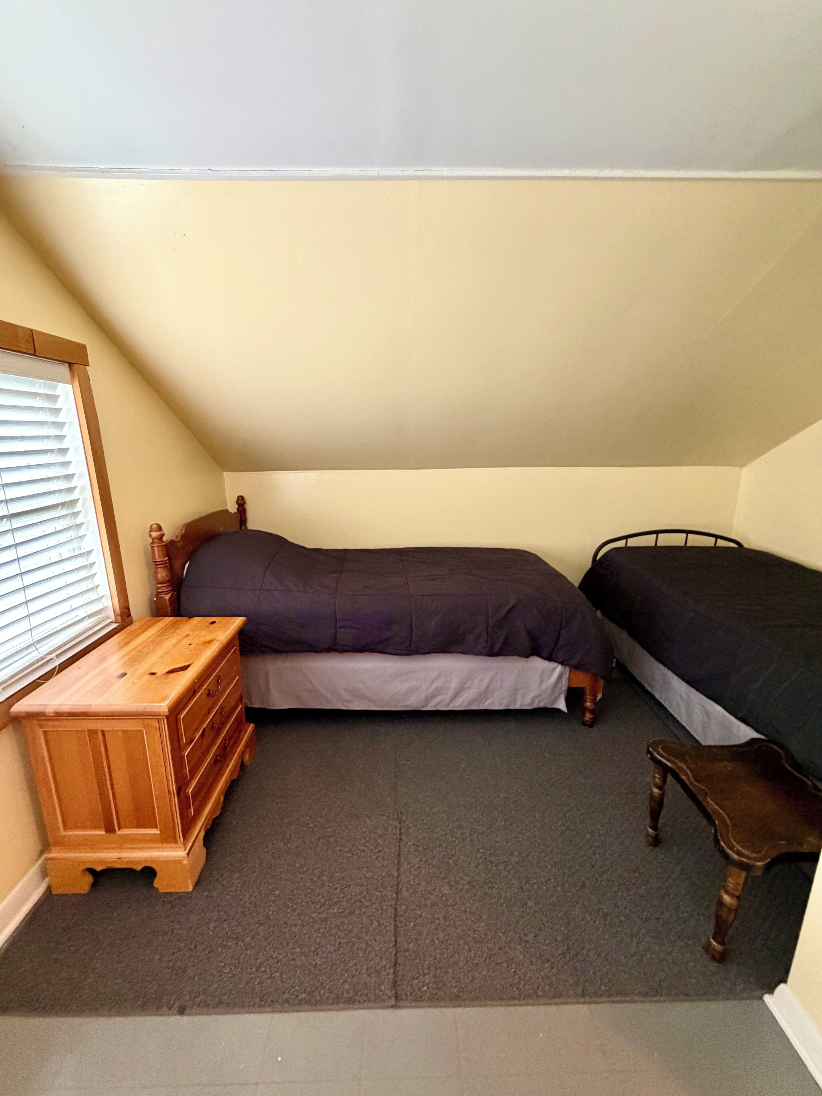 Une petite pièce triangulaire, avec un lit simple, une commode en bois et une fenêtre équipée de stores.
