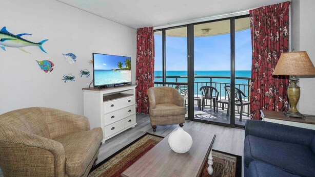 A room with a TV, a sofa, a chair, a table, and a view of the ocean through large windows.