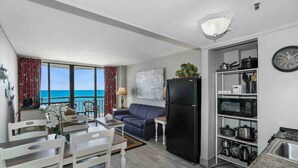 A compact living space with a dining area, a sofa, a black refrigerator, and a kitchen area with shelves and a sink.