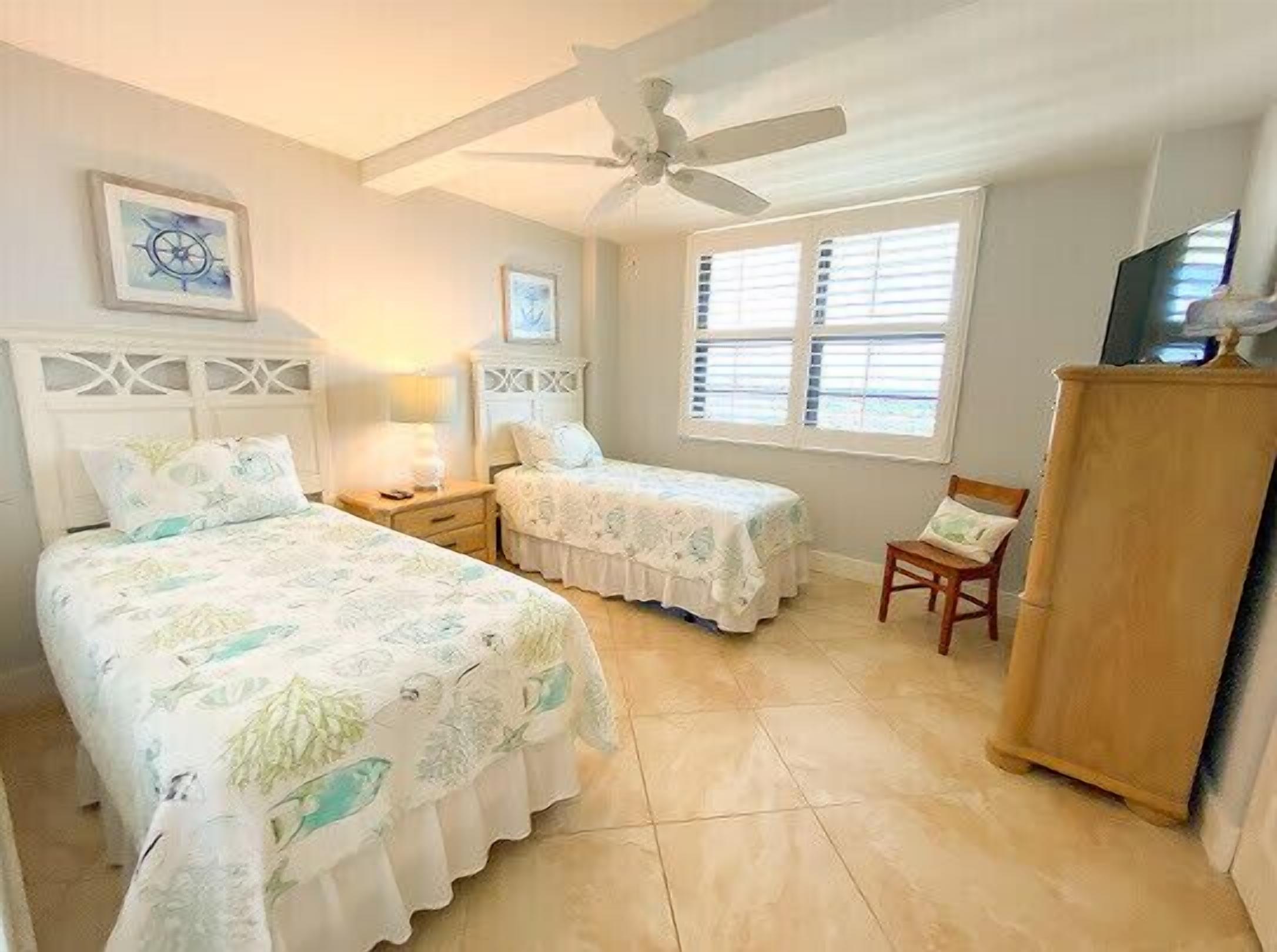 A bedroom with two beds, a ceiling fan, a window with blinds, and a wooden cabinet.