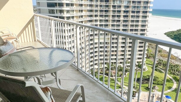 Ein Balkon mit Glastisch und zwei Stühlen, mit Blick auf ein Strandhotel mit mehreren Balkonen und freier Meerblick.