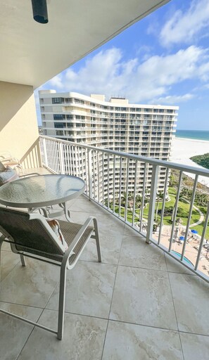 A balcony with a glass table and two chairs, overlooking a beachfront hotel with multiple balconies and a clear view of the ocean. - Marco Updated Beach Front Condo, 2Br 2Ba, 1605 - T4, Pool, Beach & Ocean Views (Marco Island)