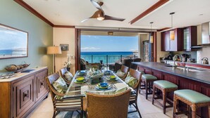 A dining area with a view of the ocean, a ceiling fan, and a kitchen with wooden cabinets.