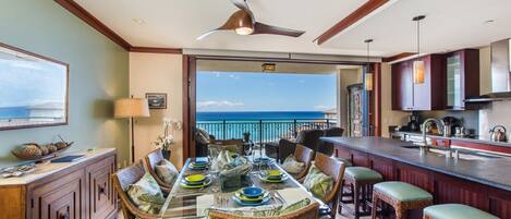 A dining area with a view of the ocean, a ceiling fan, and a kitchen with wooden cabinets.
