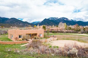A single-story adobe house with a flat roof and a front porch, surrounded by a fenced yard and a mountainous backdrop.