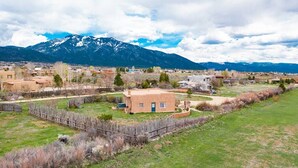 A rural landscape with a small house surrounded by a fenced yard, a mountain range in the background, and a clear sky.