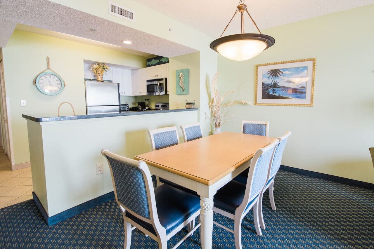 A dining area with a wooden table and blue upholstered chairs, a kitchen with white cabinets, a clock, and a framed picture on the wall.
