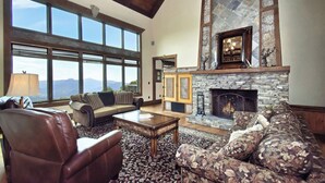 A spacious living room with a stone fireplace, large windows offering mountain views, leather furniture, and a high ceiling with exposed beams. - Multi-Million Dollar Mountain Estate-FOUR STATE VIEWS (Banner Elk)