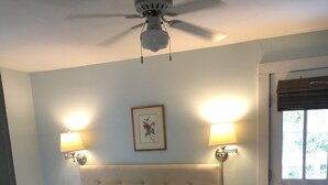Un dormitorio con cama, dos lámparas de noche, un ventilador de techo, un cuadro enmarcado y una puerta con ventanal.