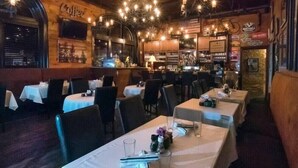 A restaurant with multiple tables, each set with white tablecloths and chairs. The interior features exposed wooden beams, hanging light fixtures, and a bar area in the background.