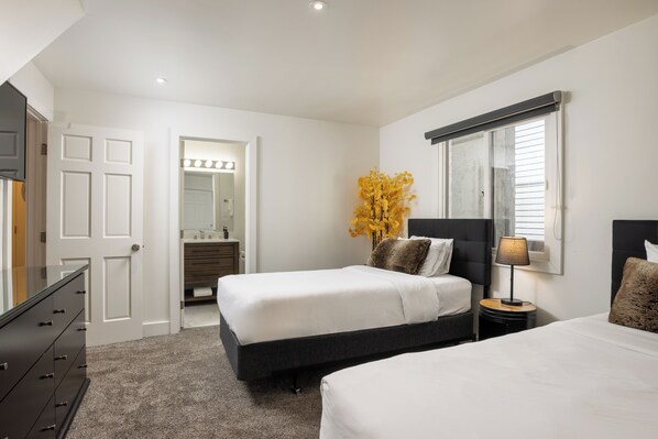 A bedroom with two beds, a dresser, a vanity with a mirror, and a window with blinds.